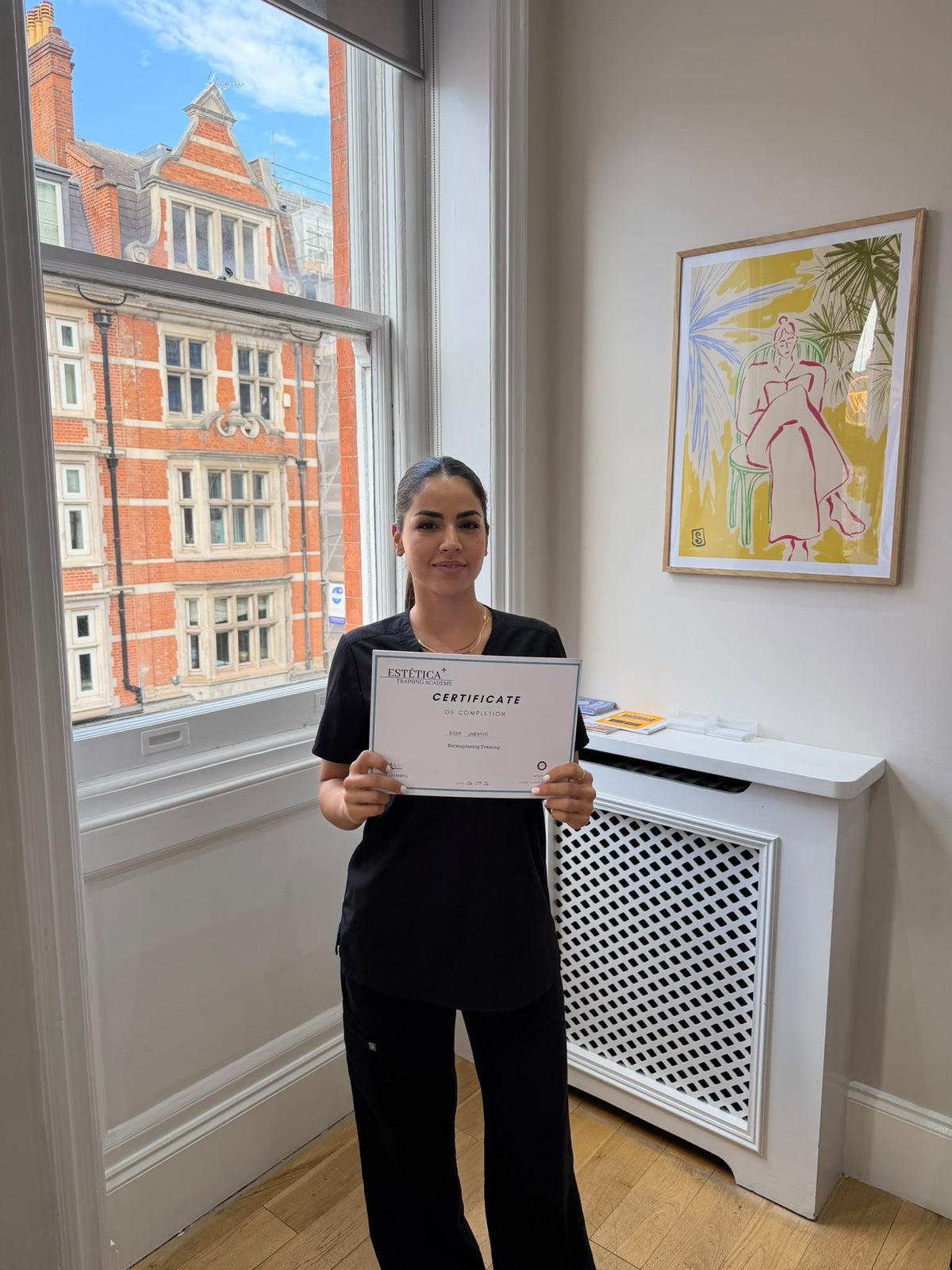 A woman in black scrubs stands in a bright room holding a certificate. A large window behind her shows a red brick building outside, and a framed colorful artwork hangs on the wall to the right.