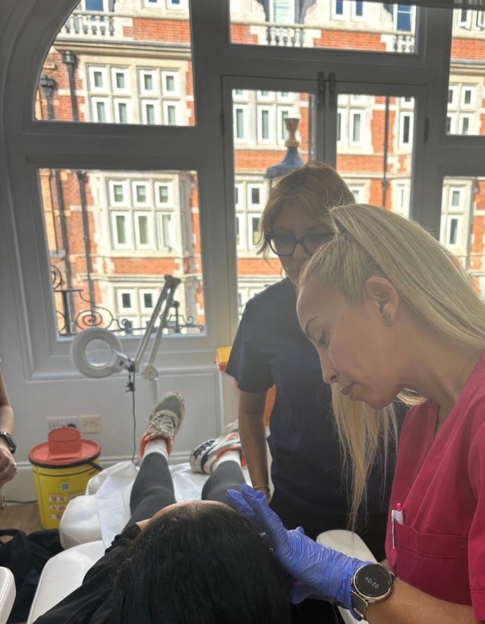 A lifestyle scene in a clinic showing a blonde woman in a pink scrub top and blue gloves performing a head massage or treatment on a client lying down, with another woman observing in the background near large windows with a brick building view injecting botox