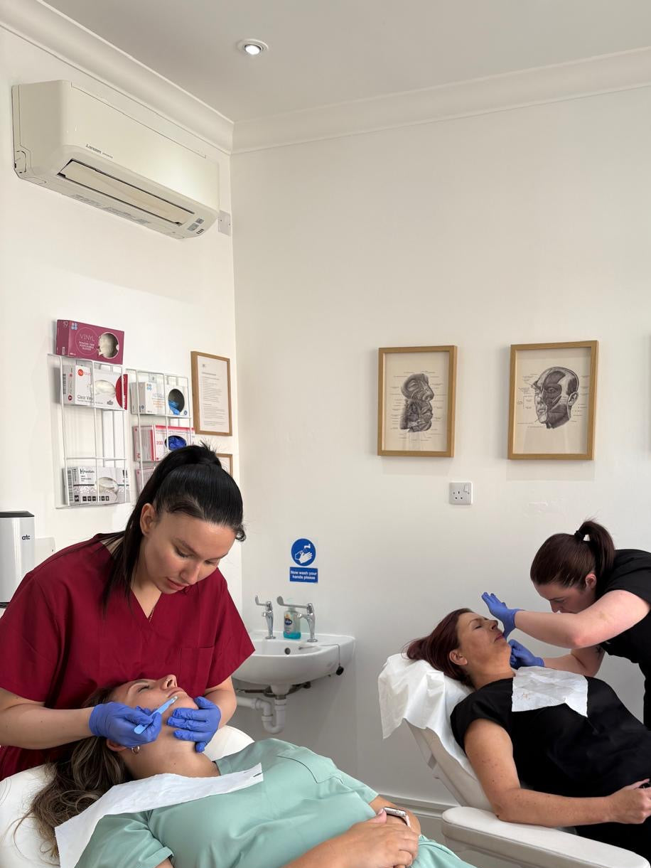 A lifestyle scene in a treatment room showing two therapists treating a client lying on a white treatment bed. One therapist in a red scrub top and blue gloves is applying a treatment to the client's face, while another therapist in a black scrub top attends to the client's abdomen.