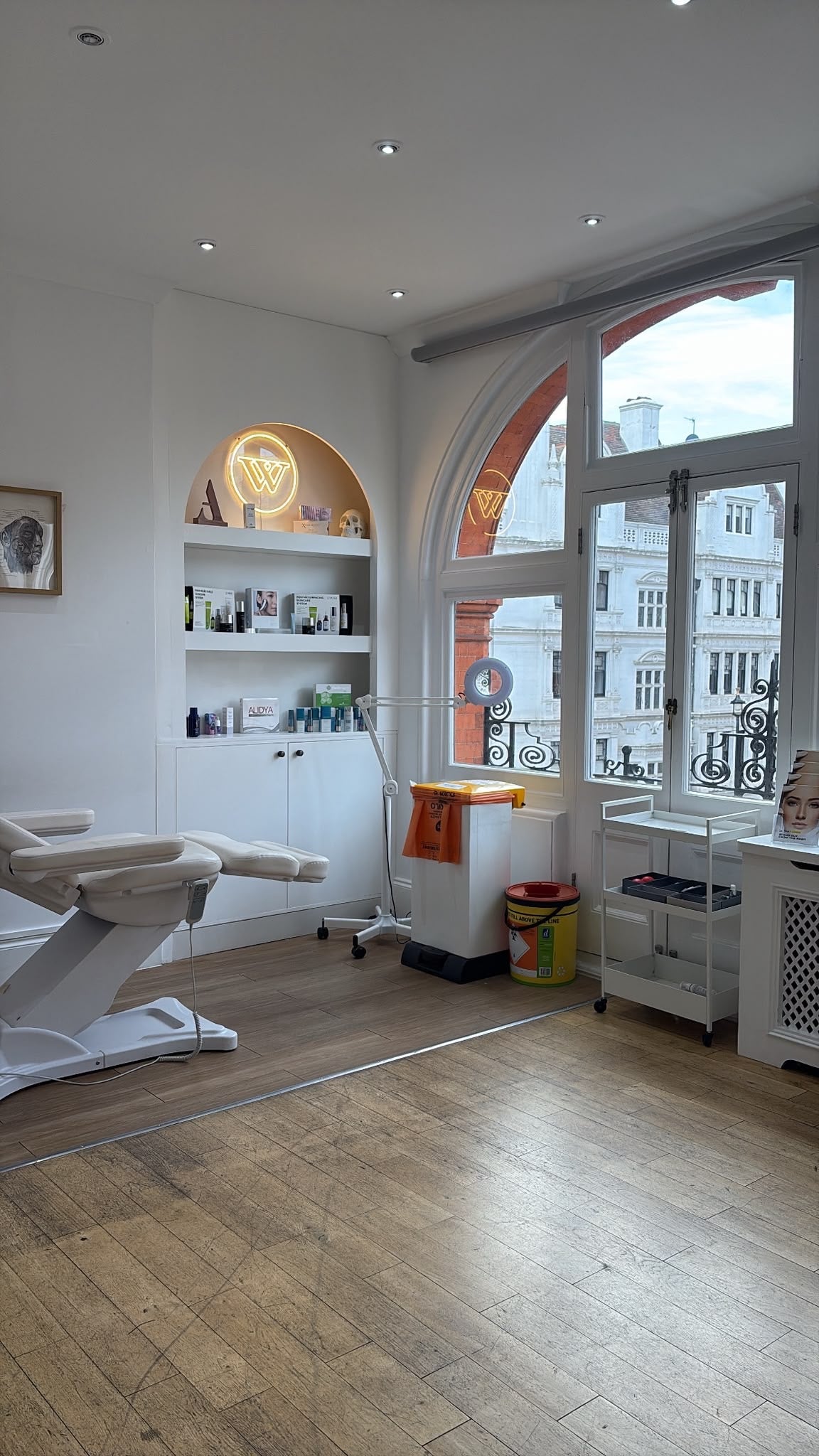 A bright, modern treatment room with a white massage table, white shelving, a large arched window, and light wood flooring.