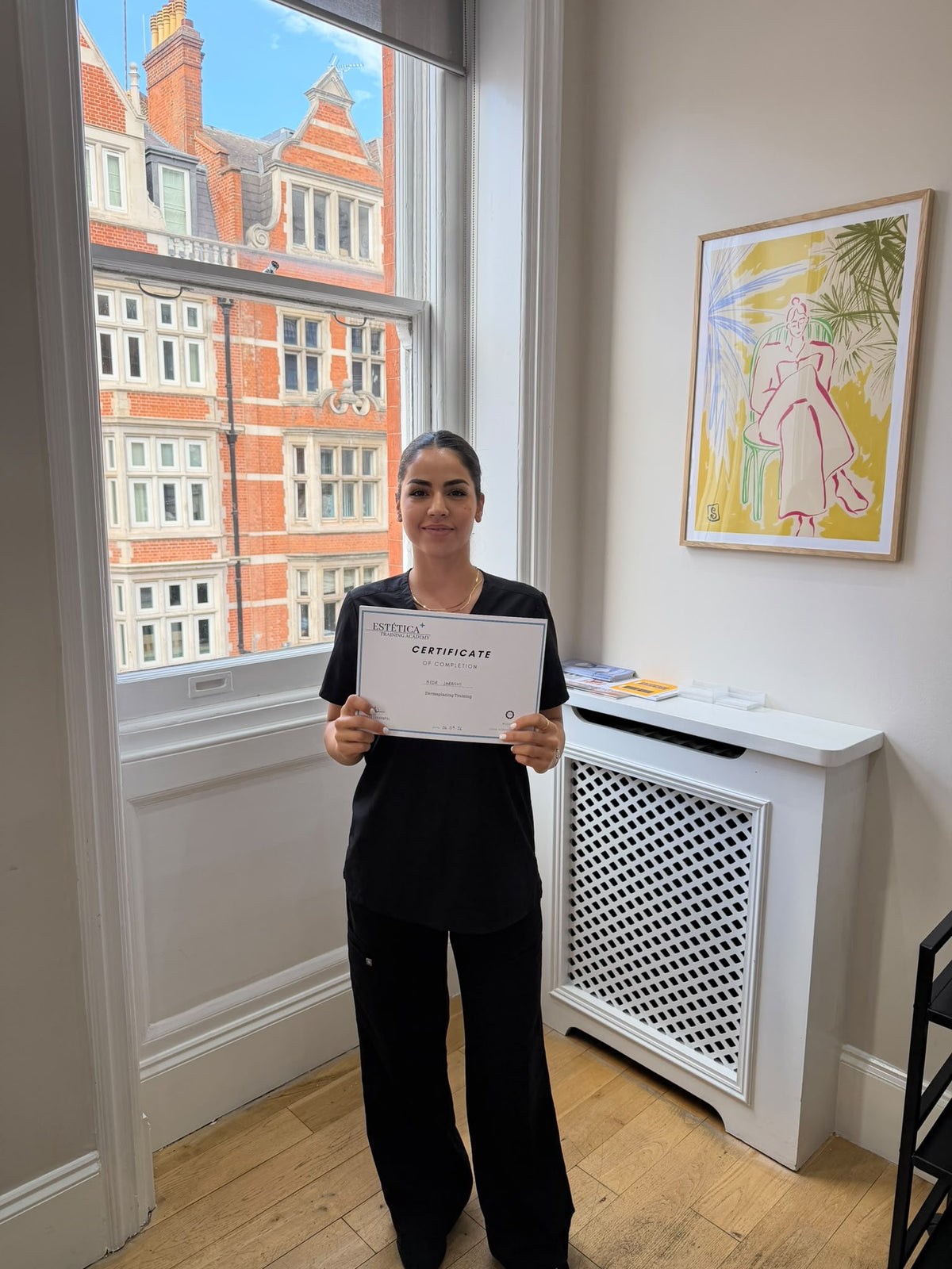 A woman in a black outfit stands in a bright room holding a certificate. A large window behind her shows red brick buildings outside, and a framed painting hangs on the wall above a white cabinet.