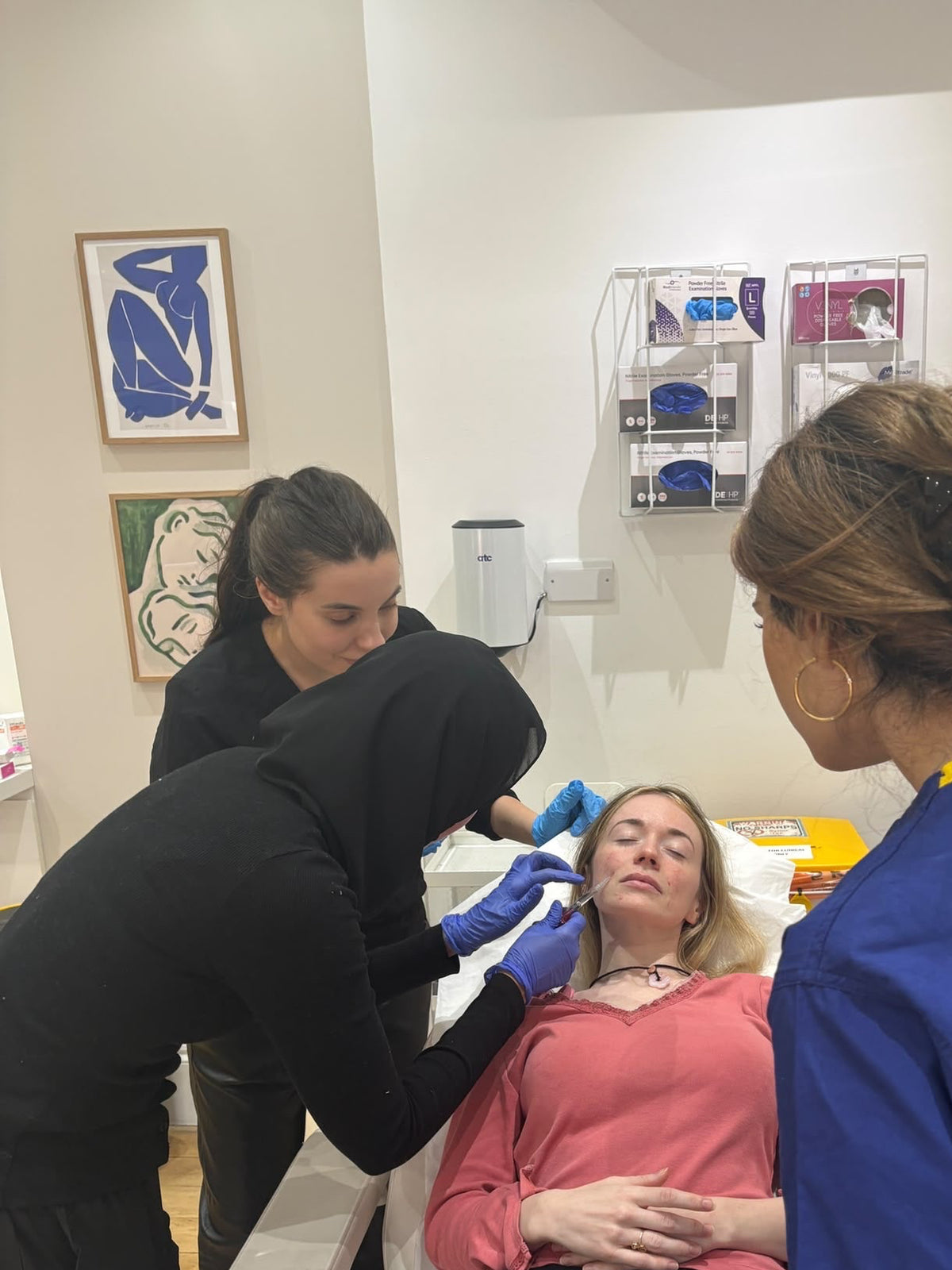 A woman in a pink top lies on a treatment bed while two staff members attend to her in a clinical room. One staff member wears a black hoodie and blue gloves, and the other wears blue scrubs.