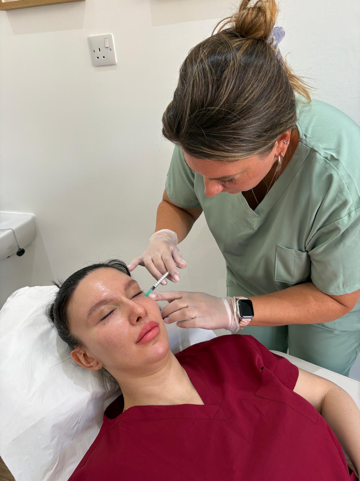 A cosmetic treatment is being administered to a woman lying down, with a practitioner in sage green scrubs leaning over her. The scene takes place in a clinical room with a white treatment bed and wall.